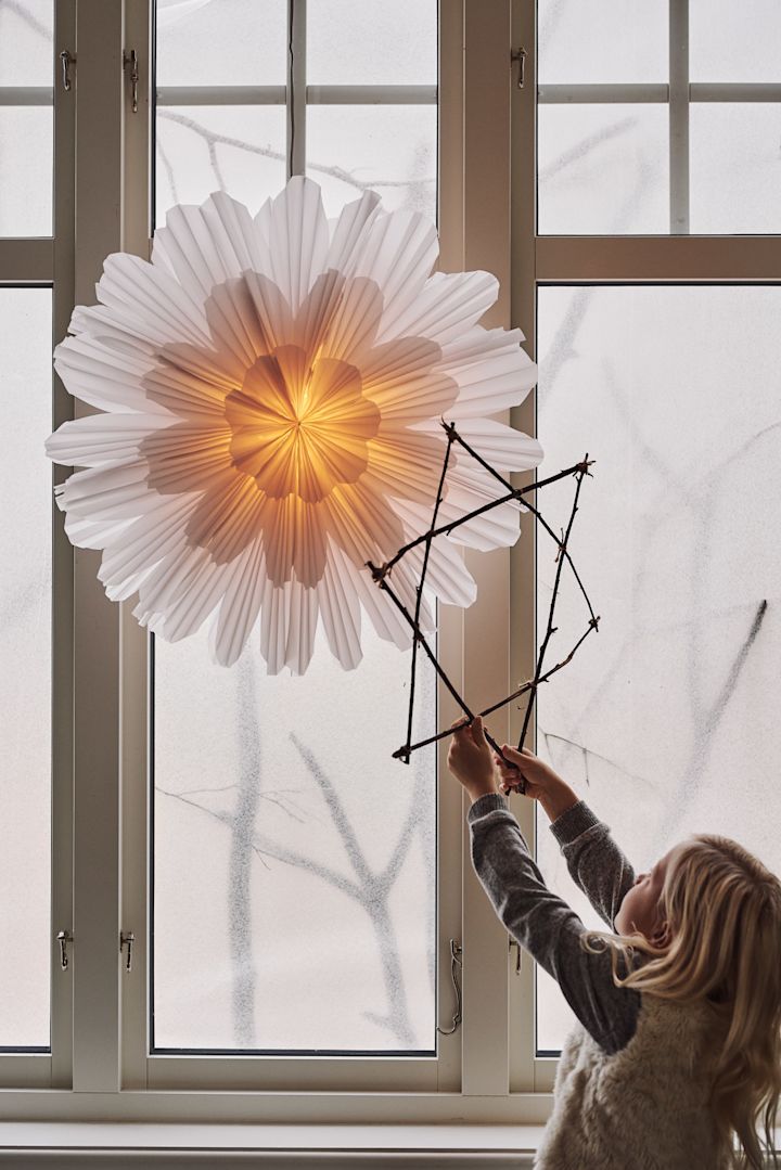 A girl holds up a homemade Christmas star out of twigs to a snöblomma paper star lantern hanging in a window. 